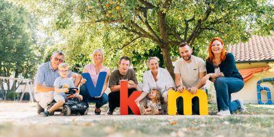 VKM Mitarbeiter, Klienten und Kitakinder posieren vor einem Baum und halten VKM Buchstaben aus Acrylglas.